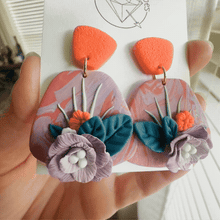 Load image into Gallery viewer, A hand holding a pair of earrings with colorful floral designs.