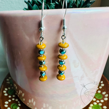 Load image into Gallery viewer, A pair of earrings with yellow beads and blue beads hanging from them, placed on a pink surface.