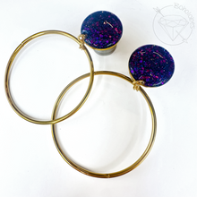 Load image into Gallery viewer, two gold hoop earrings with purple glitter accents, placed on a white background.