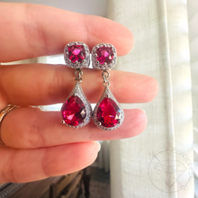 Load image into Gallery viewer, A hand holding a pair of red gemstone earrings with a sparkling crystal design.